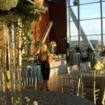 Elegant wedding reception setup featuring a woman arranging floral centerpieces in a beautifully decorated venue with large windows, showcasing tables with fine linens and chairs, emphasizing wedding rental services by Party Time Rental and Events.