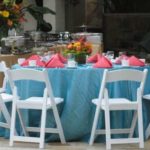 White garden chairs arranged around a decorated table with a turquoise tablecloth and pink napkins, ideal for weddings and events, showcasing rental options for Party Time Rental and Events.