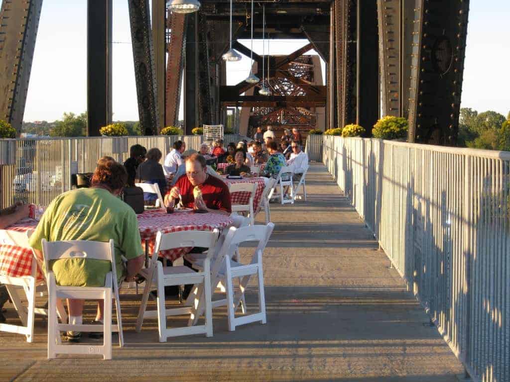 Outdoor dining event on a bridge, featuring tables with red-checkered tablecloths, guests enjoying food, and decorative flowers, associated with the Bridge to the 21st Century Dedication at the William J Clinton Presidential Library.