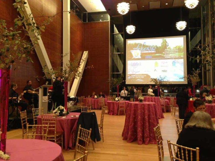 Event setup at the Clinton Presidential Center for the 31st Annual Travel South Reception, featuring round tables with pink tablecloths, gold chairs, floral centerpieces, and a large screen displaying event information.