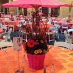 Colorful table setting featuring a vibrant pink umbrella, floral centerpiece, and orange kaleidoscope linen, designed for events by Party Time Rentals and Events.